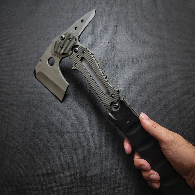 Puede incluir: Un tomahawk t&aacute;ctico con una hoja gris y un mango negro. El tomahawk presenta una cabeza de hacha afilada, una punta y un agarre texturizado para un manejo seguro. El dise&ntilde;o incluye m&uacute;ltiples tornillos y un agujero cerca de la cabeza del hacha.