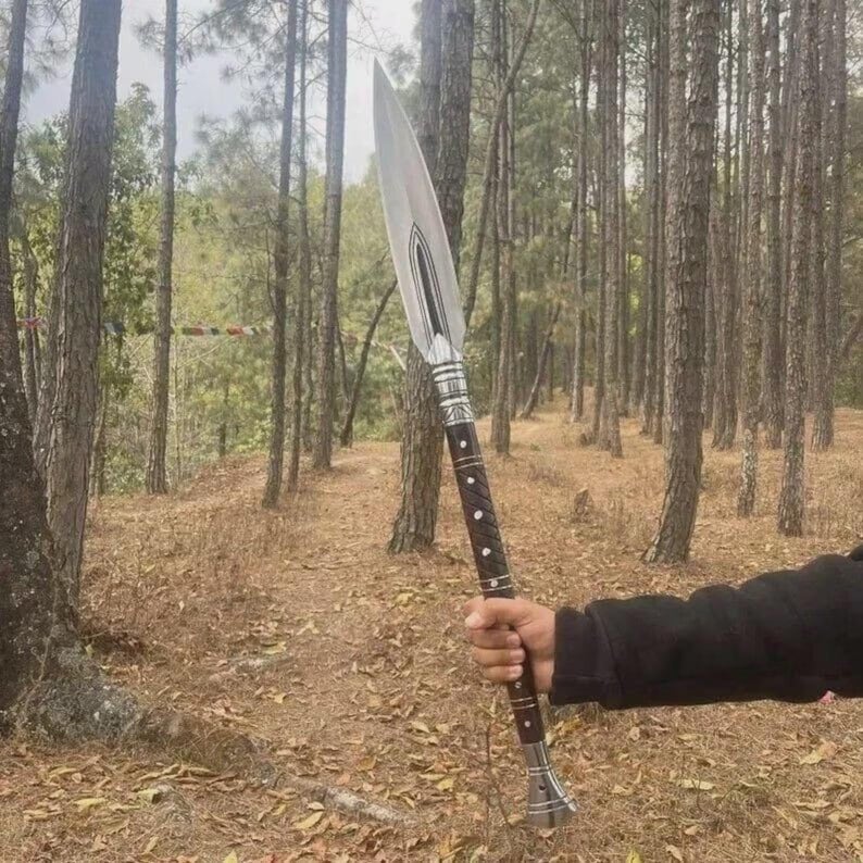 May include: A spear with a polished silver blade and a dark brown wooden handle. The handle has silver accents and white dot inlays. The spear is held in front of a forest background.