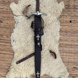 May include: A medieval-style sword with a brown leather-wrapped hilt and a circular pommel featuring a red cross emblem. The sword is housed in a black leather scabbard adorned with two white cross inlays and a leather strap with metal studs.