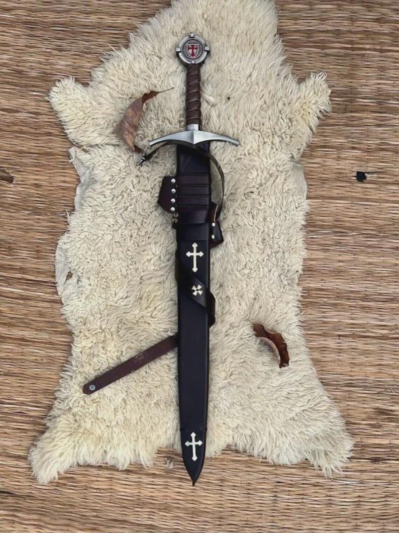 May include: A medieval-style sword with a brown leather-wrapped hilt and a circular pommel featuring a red cross emblem. The sword is housed in a black leather scabbard adorned with two white cross inlays and a leather strap with metal studs.