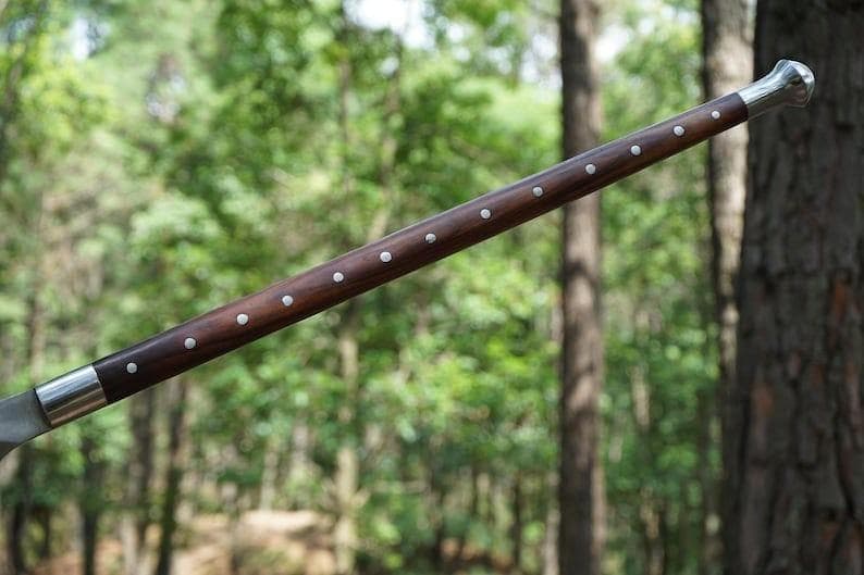 K&ouml;nnte beinhalten: Ein Gehstock aus dunkelbraunem Holz mit silbernen Nieten. Die Enden sind aus silberfarbenem Metall. Der Hintergrund ist eine verschwommene Waldszene.