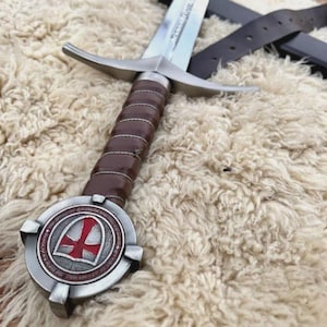 May include: A metal sword featuring a brown leather-wrapped grip and a silver crossguard. The pommel is a circular metal disc displaying a red cross emblem on a white shield background. A dark brown leather scabbard strap lies partially visible nearby.