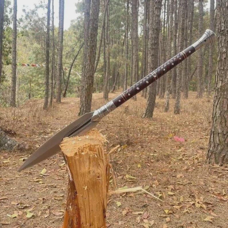 May include: A spear with a long, sharp, silver blade and a dark brown wooden shaft. The shaft is decorated with silver bands and small, dark studs. The spear is resting on a wooden stump in a forest setting.