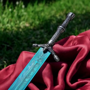 May include: A detailed close-up of a sword with a silver handle and crossguard. The blade is a vibrant blue with an intricate pattern. The sword rests on a deep red fabric against a backdrop of green grass.