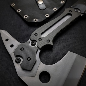 Puede incluir: Un tomahawk t&aacute;ctico con una hoja gris y un mango gris oscuro texturizado. La cabeza del hacha presenta una hoja afilada y una punta. Una funda de cuero negro con remaches es visible en el fondo.
