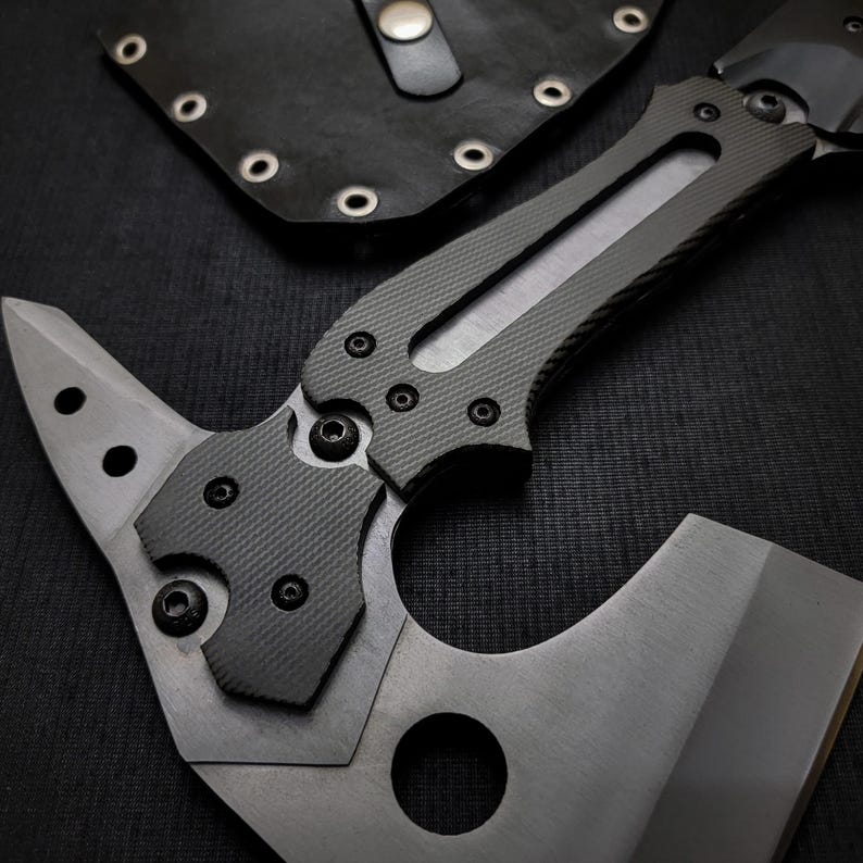 Puede incluir: Un tomahawk t&aacute;ctico con una hoja gris y un mango gris oscuro texturizado. La cabeza del hacha presenta una hoja afilada y una punta. Una funda de cuero negro con remaches es visible en el fondo.