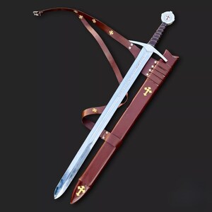 May include: A medieval sword with a silver blade and a brown leather scabbard. The hilt features a white pommel with a red cross. The scabbard and leather strap are adorned with gold crosses. The sword is approximately 40 inches long.