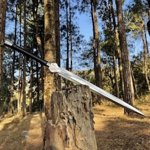 Pode incluir: Uma espada longa de dois gumes com ponta afiada, uma lâmina de metal prateado com um sulco central e um cabo preto. A espada está posicionada na diagonal, apoiada no topo de um toco de árvore vertical, rugoso e texturizado.