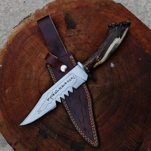 Peut inclure: Un couteau de chasse artisanal avec une lame dentelée et un manche en bois de cerf. Le couteau est dans un étui en cuir marron avec une sangle et un bouton argenté. La lame a du texte gravé.