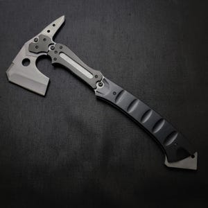 Puede incluir: Un hacha t&aacute;ctica tomahawk con mango negro y hoja gris. El hacha presenta una punta en la parte superior y una hoja curva para cortar. El dise&ntilde;o incluye un agujero cerca de la hoja, sobre un fondo oscuro.