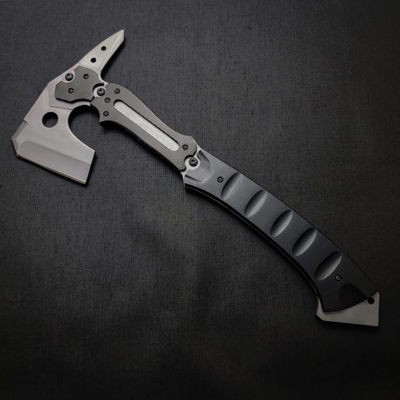 Puede incluir: Un hacha t&aacute;ctica tomahawk con mango negro y hoja gris. El hacha presenta una punta en la parte superior y una hoja curva para cortar. El dise&ntilde;o incluye un agujero cerca de la hoja, sobre un fondo oscuro.