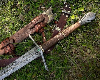 May include: A detailed overhead shot of a sword with a patterned blade and ornate wooden handle, resting on green grass. The sword is accompanied by a brown leather sheath and belt, showcasing intricate carvings and metal accents. A small green gem is set in the hilt.