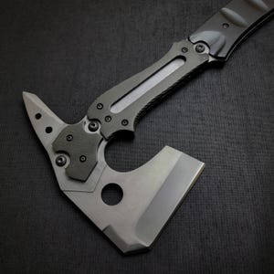 Puede incluir: Un hacha t&aacute;ctica tipo tomahawk con acabado gris. El hacha presenta una hoja afilada, un extremo puntiagudo y un mango texturizado para agarre. El dise&ntilde;o incluye un agujero cerca de la hoja y m&uacute;ltiples detalles de tornillos.