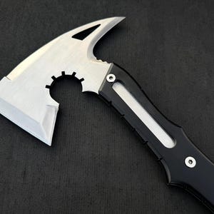 Peut inclure: Une hache tactique argent&eacute;e et noire avec une lame tranchante et un bord dentel&eacute;. La t&ecirc;te de la hache a un design unique avec un dessus incurv&eacute; et une d&eacute;coupe circulaire. Le manche est noir avec un accent blanc.