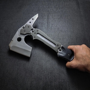 Puede incluir: Un hacha t&aacute;ctica tipo tomahawk con un dise&ntilde;o gris y negro. El hacha presenta una hoja afilada, una punta y un mango texturizado. El hacha es sostenida por una mano sobre un fondo oscuro.