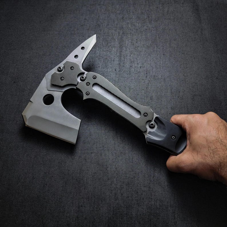 Puede incluir: Un hacha t&aacute;ctica tipo tomahawk con un dise&ntilde;o gris y negro. El hacha presenta una hoja afilada, una punta y un mango texturizado. El hacha es sostenida por una mano sobre un fondo oscuro.