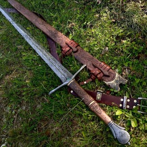 Pode incluir: Imagem detalhada de uma espada medieval com uma l&acirc;mina e punho ornamentados e estampados. A espada repousa sobre a grama verde ao lado de sua bainha de couro marrom, tamb&eacute;m intrincadamente esculpida. Uma cinta de couro marrom tamb&eacute;m &eacute; vis&iacute;vel.