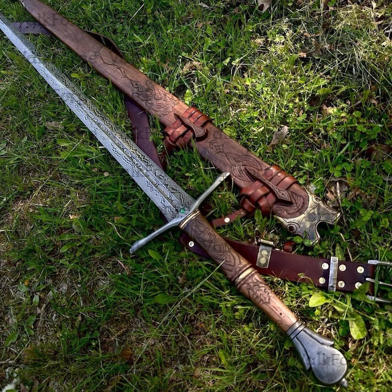 Pode incluir: Imagem detalhada de uma espada medieval com uma l&acirc;mina e punho ornamentados e estampados. A espada repousa sobre a grama verde ao lado de sua bainha de couro marrom, tamb&eacute;m intrincadamente esculpida. Uma cinta de couro marrom tamb&eacute;m &eacute; vis&iacute;vel.