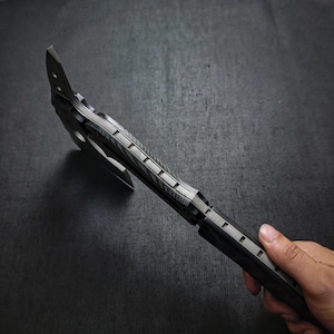 Puede incluir: Un hacha negra y plateada con un mango texturizado y una hoja afilada. El hacha tiene un agujero circular cerca de la hoja. El hacha es de metal y tiene un dise&ntilde;o moderno. El hacha est&aacute; sujeta por una mano.