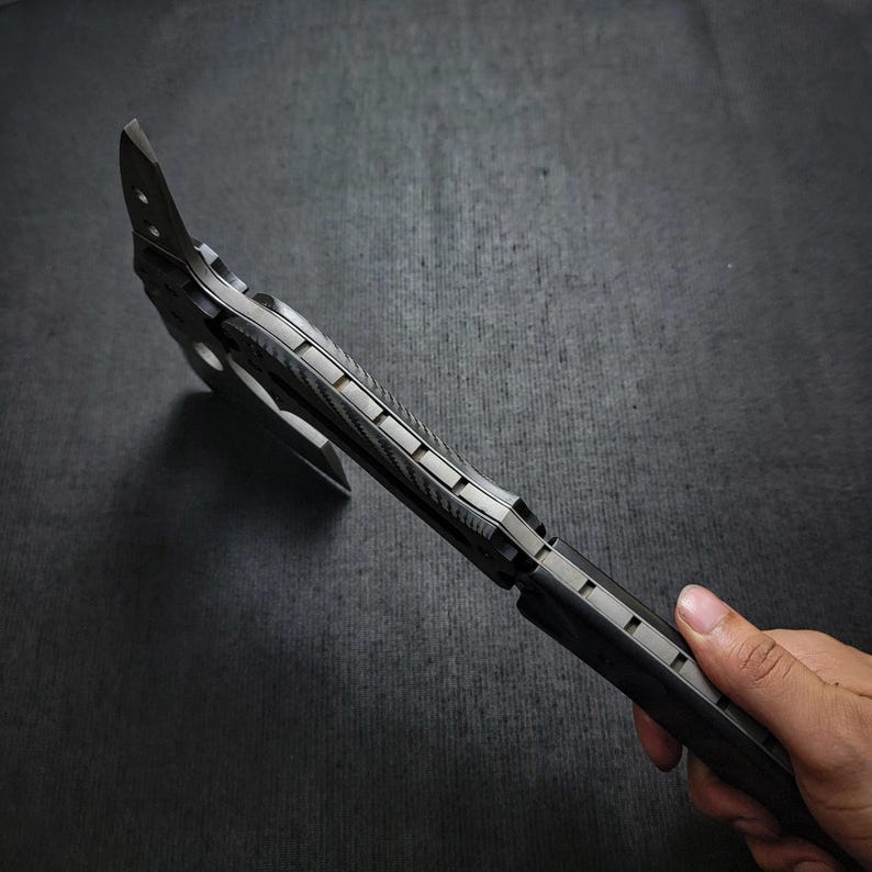 Puede incluir: Un hacha negra y plateada con un mango texturizado y una hoja afilada. El hacha tiene un agujero circular cerca de la hoja. El hacha es de metal y tiene un dise&ntilde;o moderno. El hacha est&aacute; sujeta por una mano.