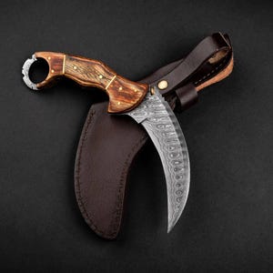May include: A curved karambit knife with a patterned steel blade and a wooden handle. The handle is accented with brass pins. The knife rests in a brown leather sheath, with a brown leather strap. The background is black.