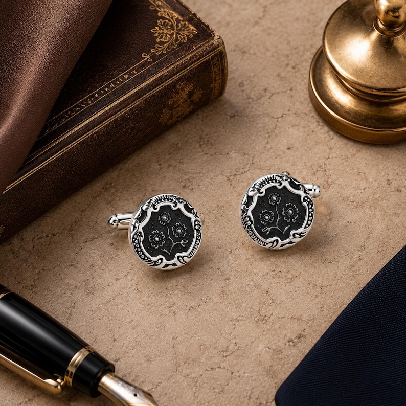 May include: A pair of round cufflinks featuring a silver-toned metal border with an intricate embossed pattern surrounding a circular black center. The black inlay displays a delicate, etched floral design with three small flowers on thin stems.