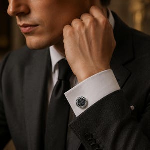 May include: A round silver cufflink featuring a dark, textured center with intricate floral-inspired engravings and a polished metallic border, secured on the white cuff of a long-sleeved shirt worn under a dark grey suit jacket.