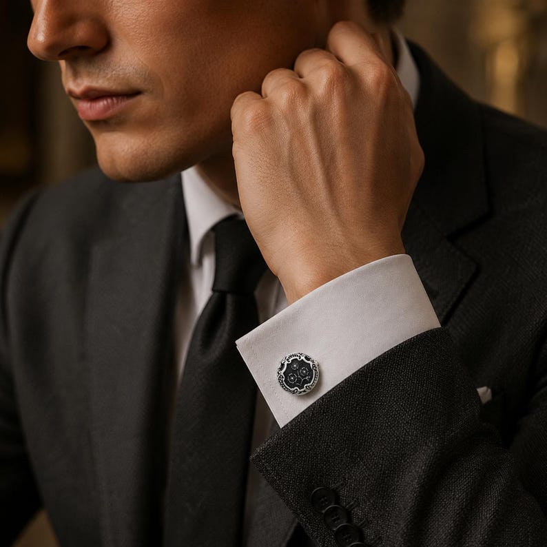 May include: A round silver cufflink featuring a dark, textured center with intricate floral-inspired engravings and a polished metallic border, secured on the white cuff of a long-sleeved shirt worn under a dark grey suit jacket.