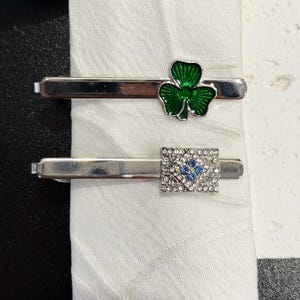 May include: Two silver-toned tie clips. One features a green shamrock design, the other a square with blue and clear crystal accents. The clips are displayed on a white fabric.