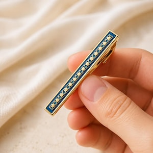 May include: A gold-toned metal tie clip featuring a rectangular shape with a central inlay of deep blue enamel. The design includes a repeating pattern of small gold circles arranged in a line down the centre, framed by a thin gold border.