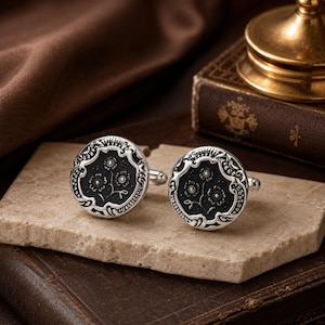 May include: A pair of round silver cufflinks featuring a black center inlay with an embossed floral design. The silver border displays intricate scrollwork patterns surrounding the dark circular face, with a standard toggle mechanism on the back.