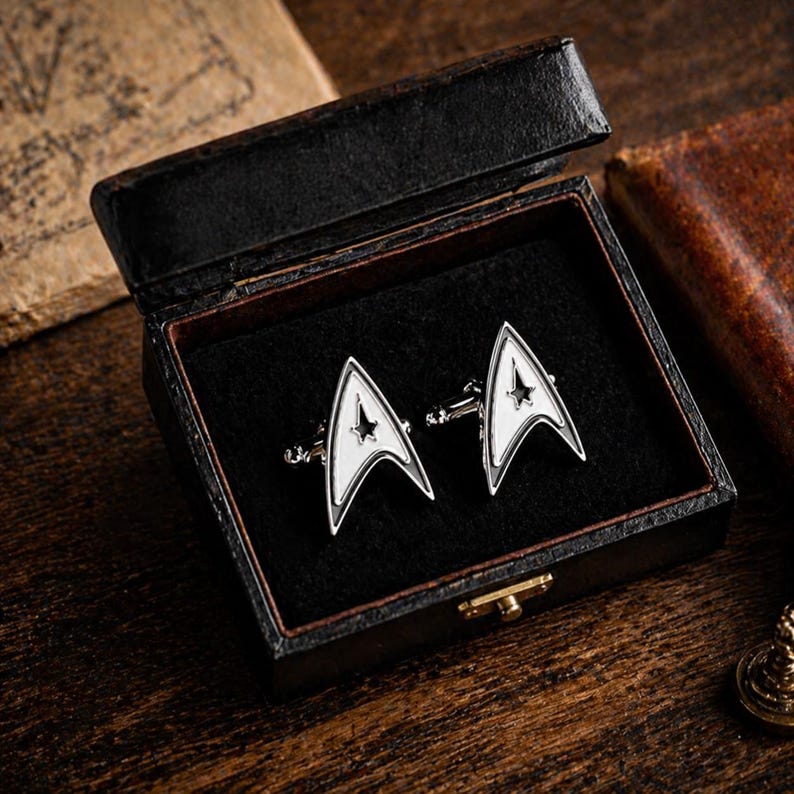 Op de afbeelding: Een paar zilverkleurige manchetknopen met een wit Starfleet-insignia-ontwerp, gepresenteerd in een zwarte kunstleren doos met een scharnierend deksel. De manchetknopen hebben een ster in het insigne.