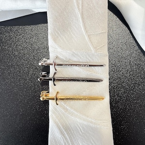 Può includere: Tre fermacravatte a forma di spada in tonalità argento e oro, esposti su una cravatta bianca fantasia. Le clip hanno elsi e lame dettagliate, aggiungendo un tocco di eleganza all'abbigliamento formale. La cravatta è bianca con un motivo sottile.