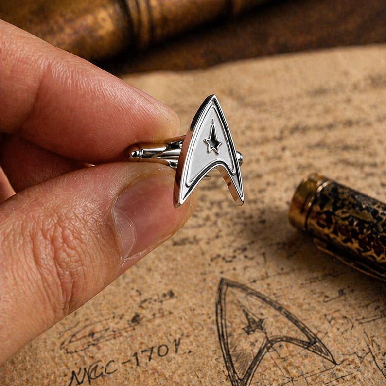 Op de afbeelding: Zilveren manchetknopen met een wit en zwart Starfleet insigne ontwerp. De manchetknopen hebben de vorm van een driehoek met een ster in het midden. Op een bruin oppervlak met de tekst "NCC-1701".
