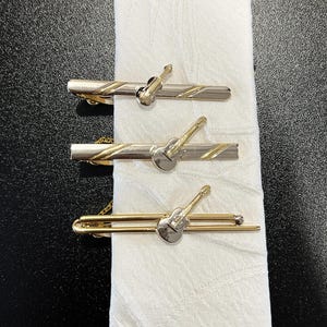 May include: Three vintage tie clips with a guitar design. The clips are silver and gold-toned, with a small guitar charm in the center. The tie clips are displayed on a white cloth.