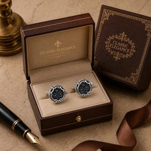 May include: A pair of round silver cufflinks with black circular inlays featuring a white floral design, presented in a brown hinged box with the text CLASSIC ELEGANCE TIMELESS STYLE printed in gold on the interior lid and the box exterior.