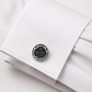 May include: A circular silver-toned cufflink with a black recessed center featuring a detailed floral engraving. The cufflink is fastened through the double-layered cuff of a crisp white dress shirt with a visible diagonal twill fabric texture.