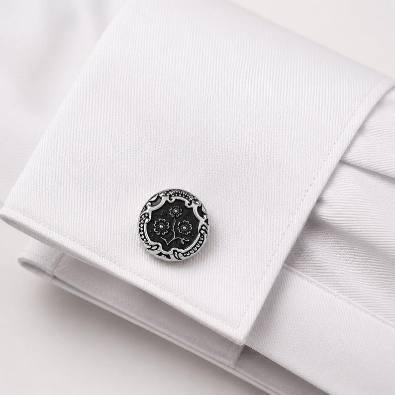 May include: A circular silver-toned cufflink with a black recessed center featuring a detailed floral engraving. The cufflink is fastened through the double-layered cuff of a crisp white dress shirt with a visible diagonal twill fabric texture.