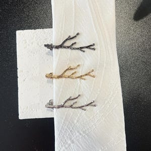May include: Three decorative napkin rings shaped like tree branches. One is black, one is gold, and one is silver. The napkin rings are clipped onto a white cloth napkin.