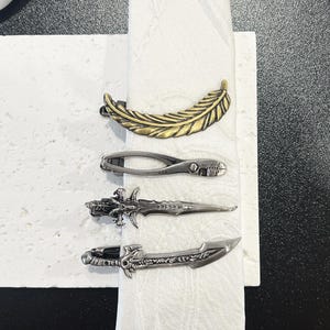 Może przedstawiać: Cztery dekoracyjne pierścienie do serwetek. Górny pierścień to złota pióro. Poniżej znajduje się srebrny pierścień z nożycowatym wzorem. Dwa dolne pierścienie to srebrne wzory sztyletów.