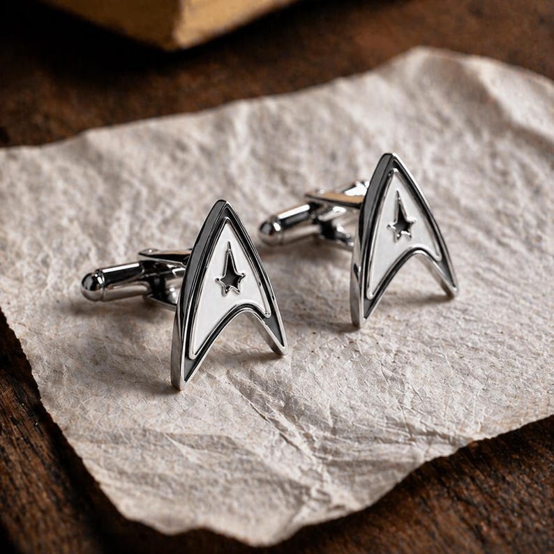 Op de afbeelding: Een paar zilverkleurige manchetknopen met een wit en zwart Starfleet-insignet ontwerp. De manchetknopen hebben een gepolijste afwerking en worden gepresenteerd op een getextureerd, gebroken witte ondergrond.