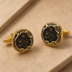 May include: A pair of round cufflinks featuring a polished gold-tone metal border with an intricate embossed pattern surrounding a recessed black circular center. The black center displays a detailed floral motif with three small flowers on stems.