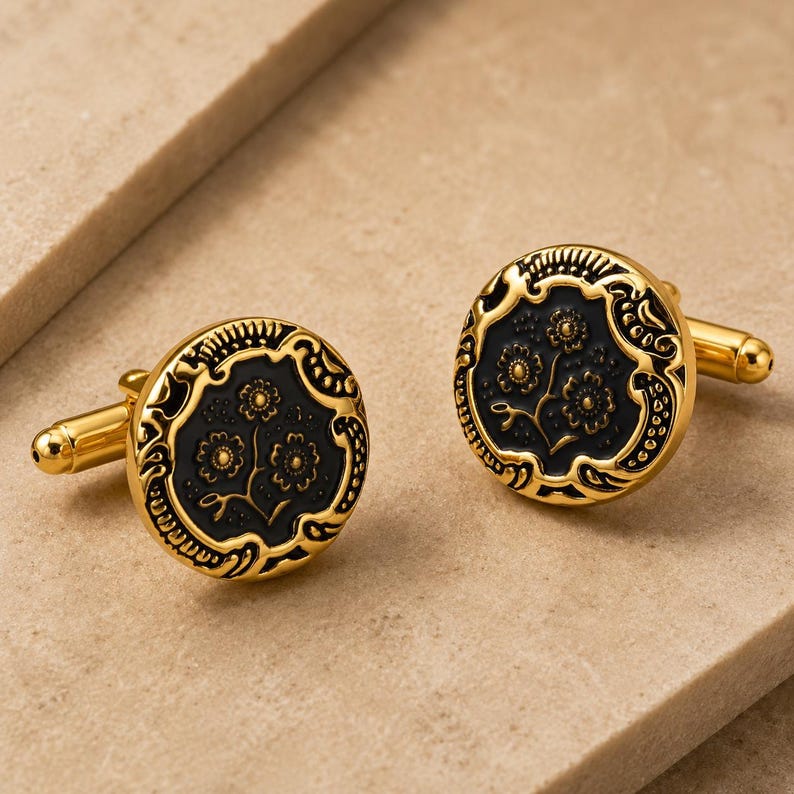 May include: A pair of round cufflinks featuring a polished gold-tone metal border with an intricate embossed pattern surrounding a recessed black circular center. The black center displays a detailed floral motif with three small flowers on stems.