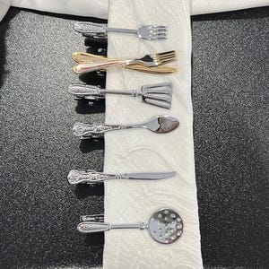 Peut inclure: Ensemble de pinces à cravate miniatures en forme de couverts, comprenant des fourchettes, une spatule, une cuillère, un couteau et une cuillère ajourée. Les pinces sont argentées et dorées, disposées sur un tissu blanc.