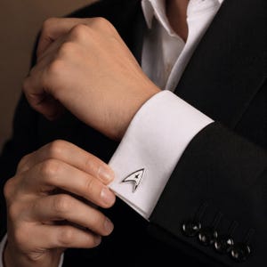 Op de afbeelding: Zilverkleurige manchetknopen met het zwart-witte Starfleet-insigne worden getoond op een witte overhemdmouw. De manchetknopen staan op een zwart colbert. De persoon past de manchetknopen aan.