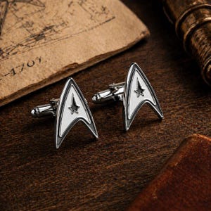 Op de afbeelding: Set zilverkleurige manchetknopen met een wit en zwart Starfleet-insigne ontwerp. De manchetknopen liggen op een donkerhouten oppervlak, met oud papier op de achtergrond, wat een vintage uitstraling cre&euml;ert.
