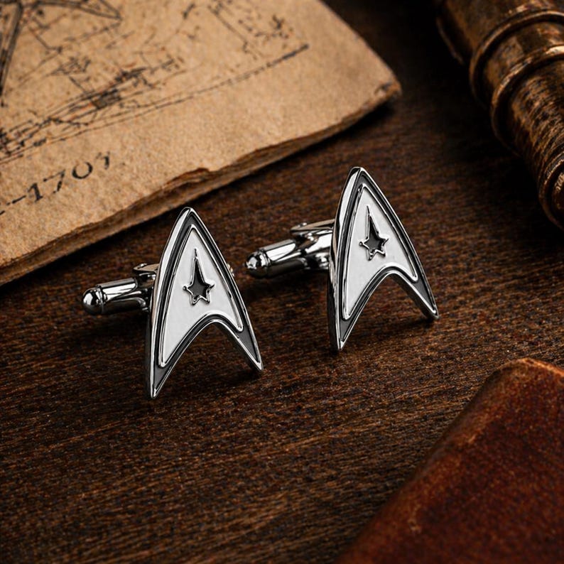 Op de afbeelding: Set zilverkleurige manchetknopen met een wit en zwart Starfleet-insigne ontwerp. De manchetknopen liggen op een donkerhouten oppervlak, met oud papier op de achtergrond, wat een vintage uitstraling cre&euml;ert.