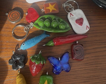 Peut inclure: Une collection de 13 breloques en argile faites à la main, comprenant une gousse de pois, un cœur rouge, une étoile, un papillon, une fraise, une baguette, un piment rouge, un poisson, une fleur, un tourbillon et un as de cœur. Les breloques sont peintes dans différentes couleurs, notamment le rouge, le vert, le bleu, le jaune et le marron.