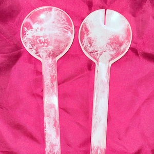 May include: A pair of clear salad servers with a white, cloud-like pattern. One is a spoon, the other a fork. The servers are on a pink background, suggesting a kitchen or dining theme.
