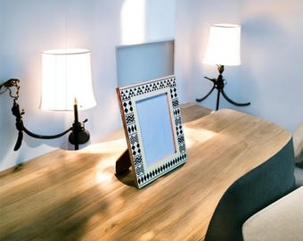 May include: A decorative wooden picture frame with a black and white geometric pattern stands on a wooden table. Two lamps with white shades are on either side of the frame. The table has a natural wood finish.